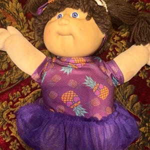 Pode incluir: Uma boneca com cabelo de lã castanho em tranças, olhos azuis e rosto redondo. A boneca veste uma blusa roxa de manga curta com um padrão repetitivo de abacaxi e uma saia de tule roxa a combinar com um laço.