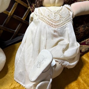 Puede incluir: Una muñeca de tela con cabello de lana marrón y ojos azules, vestida con un vestido color crema con un cuello de encaje detallado. La muñeca tiene zapatos blancos con una firma impresa en la suela, con rostro redondeado y cuerpo blando.