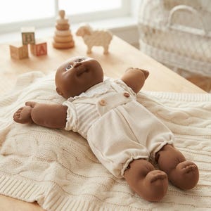 May include: A dark-skinned doll with a round face and brown eyes, wearing a cream-colored short-sleeved romper with a white lace collar and small brown buttons on the front straps, lying on a cream-colored cable-knit blanket.