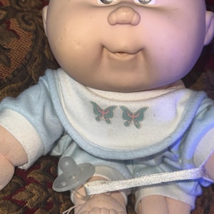 Puede incluir: Una muñeca calva con cara redonda, ojos marrones y nariz pequeña, que lleva un conjunto azul claro y blanco con un babero que presenta dos gráficos de mariposas verdes y rosas. Un chupete de plástico transparente está unido por una cinta blanca.