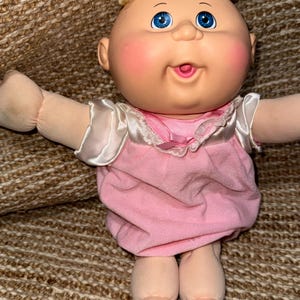 Puede incluir: Una muñeca con cabeza redonda, piel clara, mejillas sonrosadas y ojos azules brillantes. La muñeca tiene el pelo rubio rizado y lleva un vestido rosa con mangas de satén blanco, un cuello de encaje y un pequeño lazo rosa en el centro.