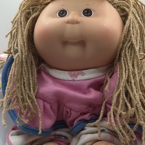 Puede incluir: Una muñeca con cara redonda de vinilo suave, ojos marrón oscuro y cabello de lana marrón claro. La muñeca viste una blusa rosa con cuello blanco con pequeños patrones rosas y pantalones blancos con un estampado colorido. Cuerpo de tela relleno.