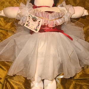 Puede incluir: Una muñeca de tela con cabello oscuro rizado, que viste un corpiño rojo metálico y una falda de tul blanco en capas. La muñeca tiene mangas con volantes, zapatillas de ballet con cintas y una etiqueta rectangular unida al corpiño.