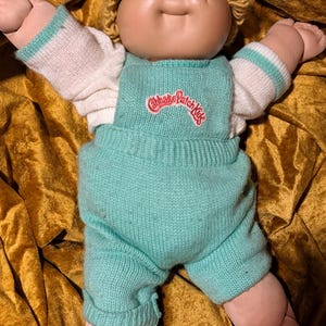 Pode incluir: Uma boneca Cabbage Patch Kids com pele clara, olhos verdes e cabelo de lã loira. A boneca veste uma camisa branca de manga comprida com punhos verde-azulados e um macacão de malha verde-azulado com o logótipo vermelho no peito.