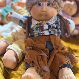 May include: A soft-sculpture doll with brown yarn hair, blue eyes, and dimpled cheeks. The doll wears a blue plastic hat, a plaid collared shirt, and brown suede-like chaps with fringe detailing over dark pants, featuring a light skin tone.