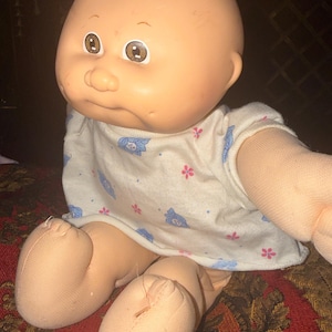 May include: A bald doll with a round, peach-toned vinyl head, brown eyes, and a soft fabric body. The doll wears a short-sleeved, off-white shirt featuring a repeating pattern of small blue shapes and tiny pink flowers across the fabric surface.