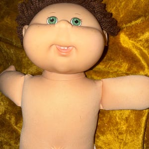 May include: A doll with a round, light-toned vinyl head, green eyes, and short, textured brown yarn hair. The doll features a soft, light-toned fabric body with a prominent belly button and a small, open mouth showing two visible front teeth.