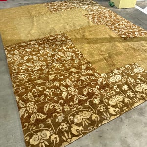 Puede incluir: Una alfombra rectangular grande con un diseño de patchwork. La alfombra presenta secciones marrones y beige lisas y estampadas. Las secciones estampadas muestran un motivo floral y de hojas en tonos crema y marrón. La alfombra está hecha de un material suave y afelpado.