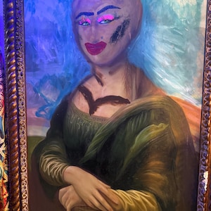 Puede incluir: Una pintura al óleo de la Mona Lisa con maquillaje exagerado, que incluye sombra de ojos rosa brillante y lápiz labial rojo. El retrato está ambientado sobre un fondo azul y enmarcado en un marco dorado adornado. La expresión del sujeto está alterada.