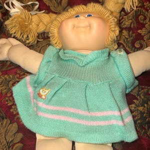 May include: A doll with a soft fabric body, light skin, and blue eyes. The doll has blonde yarn hair styled in two braided pigtails with white ribbons. It wears a mint green knitted dress featuring two horizontal pink stripes and a small duck patch.