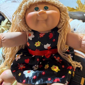 May include: A doll with a round face, green eyes, and long blonde yarn hair. The doll wears a black sleeveless dress featuring a colourful floral pattern of red, yellow, and white flowers, accented by a solid red ribbon waistband with a small fabric rose.
