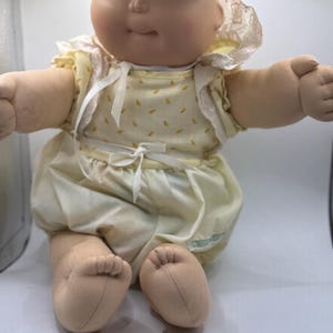 Puede incluir: Una muñeca de tela con cabeza redonda color beige, ojos azules y cabello de lana. La muñeca viste un pelele amarillo pálido con un pequeño estampado naranja, un cuello de encaje blanco y un lazo blanco en la cintura con bombachos a juego.