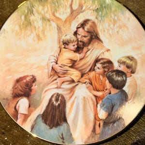 May include: A round decorative plate featuring a painted illustration of a long-haired, bearded figure in light-colored robes sitting under a tree, surrounded by six children. The scene is rendered in soft, warm tones with a gold-colored rim.