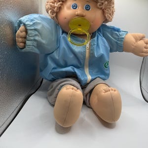 Puede incluir: Una muñeca de cuerpo blando con piel bronceada, ojos azules y cabello de lana rizado de color marrón claro. La muñeca viste una chaqueta azul claro con cremallera y un logo verde en el pecho y pantalones grises. Un chupete amarillo está en la boca.
