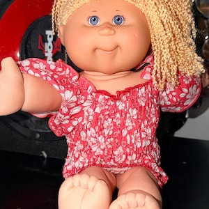 May include: A doll with light skin, blue eyes, and freckles, featuring curly blonde yarn hair styled with a small purple bow. The doll wears a red and white floral print top with ruffled sleeves and a smocked bodice, showing bare feet and arms.