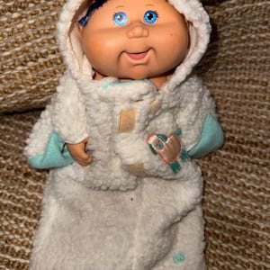 Puede incluir: Una muñeca con ojos azules y cabello oscuro que viste un mameluco con capucha de forro polar sherpa blanco con detalles en azul claro. El mameluco presenta un gráfico de oveja bordado en el pecho y un pequeño logotipo cerca del dobladillo inferior.