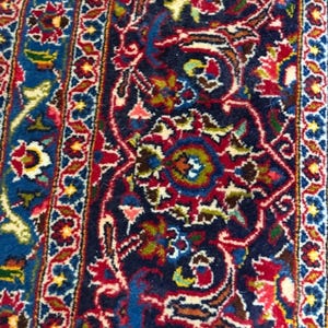 May include: Close-up of a detailed, ornate rug with a rich color palette. The rug features a deep navy blue background with intricate patterns in red, cream, yellow, and green. Floral and geometric designs create a traditional aesthetic.