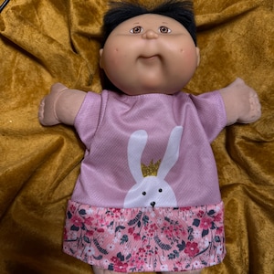 May include: A doll with a round face, dimpled cheeks, and short black hair. The doll wears a short-sleeved pink shirt featuring a white rabbit graphic with a gold crown, paired with a skirt displaying a pink, white, and dark floral pattern.