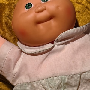Puede incluir: Una muñeca con cabeza redonda de vinilo en tono melocotón, ojos verdes y mejillas con hoyuelos. La muñeca viste una camisa blanca con cuello, mangas cortas y una falda de cuadros en verde claro y blanco. Tiene la cabeza lisa y sin pelo.
