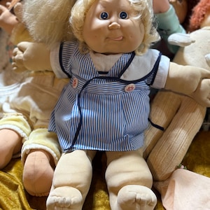 Könnte beinhalten: Eine Puppe mit blondem Haar, blauen Augen und einem runden Gesicht, bekleidet mit einem blau-weiß gestreiften Kleid mit weißem Kragen und zwei kleinen Knöpfen. Die Puppe hat einen weichen Stoffkörper, sichtbare Zehen und liegt zwischen anderen Puppen.