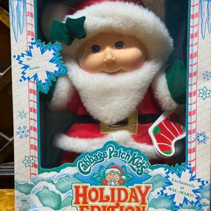 Pode incluir: Uma boneca Cabbage Patch Kids Holiday Edition vestida de Papai Noel com traje vermelho, acabamento em pele sintética branca e um detalhe de azevinho verde no gorro. A boneca está numa caixa retangular com etiqueta de presente To/From, flocos de neve e o texto Exclusively at WAL-MART.