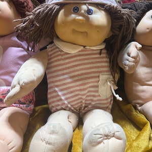 Puede incluir: Una muñeca de tela con cabello de lana marrón, ojos azules y piel clara. La muñeca viste un mameluco a rayas rosas y blancas con un bolsillo pequeño y un gorro a rayas a juego con un nudo en la parte superior. Tiene cara y extremidades redondeadas.