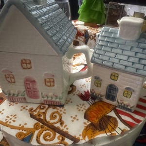 May include: Two ceramic house-shaped teapots. The larger teapot has a white body, a gray roof, and a pink door. The smaller teapot has a white body, a gray roof, and a blue door. Both have handles and are decorated with floral details.