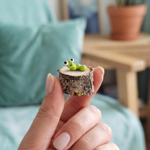 May include: A small, light green clay caterpillar figurine with large white and black eyes, perched on top of a circular, rough-textured wooden log slice with visible bark and a flat, light-colored top surface.