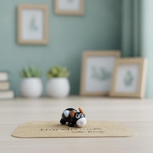 May include: A small glass figurine of a black and white cow with orange horns, positioned on a rectangular brown card with the text "Handmade with love" printed in black ink across the center of the card surface.