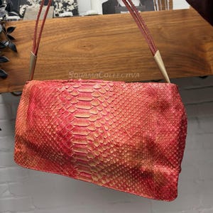 May include: A red and gold snakeskin-patterned handbag with thin, red straps and gold-coloured metal accents. The rectangular bag is held up by a person's hand. The bag's texture is visible, and the overall design is stylish.