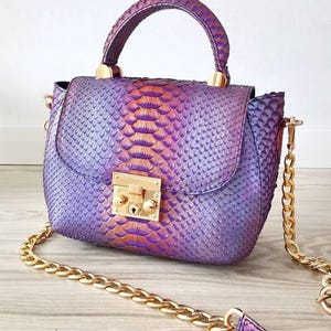Puede incluir: Bolso de mano morado y naranja con estampado de piel de serpiente, con una correa y asa de cadena dorada. El bolso tiene un cierre de broche dorado y una forma estructurada. El asa tiene un estampado de piel de serpiente a juego.