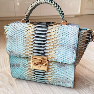 May include: A turquoise and gold snakeskin handbag with a top handle and a gold clasp. The bag features a flap closure and a detachable chain strap. The snakeskin pattern is detailed and textured, with a mix of blue, gold, and black scales.