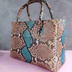 Può includere: Borsa rettangolare con motivo a pelle di serpente nei toni del marrone, beige e verde acqua. La borsa ha due manici arrotondati e inserti verde acqua sul davanti e sui lati. La borsa è realizzata in un materiale strutturato.