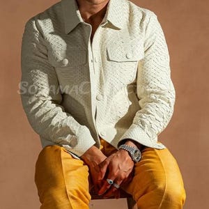 Puede incluir: Chaqueta clara y texturizada con botones y dos bolsillos en el pecho. La chaqueta se combina con pantalones amarillos brillantes. También se ve un reloj plateado y un anillo. La marca "Soamac" es visible en la chaqueta.