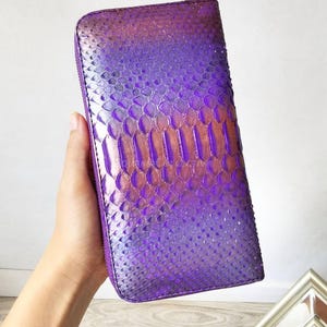 May include: A rectangular zip-around wallet featuring a textured snakeskin pattern with a vibrant gradient of purple and metallic copper tones. The wallet has a structured shape with rounded corners and a matching purple zipper along the edge.