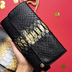 Może przedstawiać: Czarna kopertówka ze złotym łańcuszkiem. Kopertówka wykonana jest z teksturowanego czarnego materiału ze złotymi akcentami. Kopertówka jest prostokątna z klapką i srebrnym guzikiem. Tło jest czerwone ze złotym konfetti.