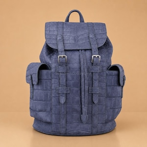 Pode incluir: Uma mochila azul com um padrão texturizado de pele de crocodilo. A bolsa inclui uma alça superior, uma aba com duas fivelas de metal prateado, um fecho de cordão e dois bolsos laterais com abas na parte externa.