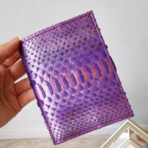 May include: A purple leather wallet with a snakeskin pattern. The wallet is rectangular and has a textured surface. The color transitions from a deep purple to a lighter pinkish hue.