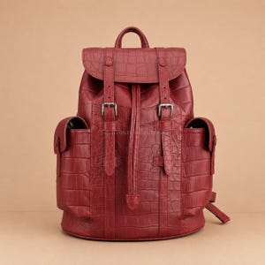 Può includere: Uno zaino rosso scuro con una texture a rilievo effetto coccodrillo. La borsa presenta un manico superiore, una patta con due cinghie verticali fissate da fibbie in metallo argentato e due tasche laterali con chiusura a patta e bottoni a pressione.
