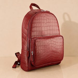 Puede incluir: Una mochila de color rojo intenso con un patrón de textura de piel de cocodrilo. La bolsa incluye un compartimento principal grande con cremallera plateada, un bolsillo frontal más pequeño, un asa superior y correas ajustables.