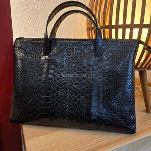 Puede incluir: Bolso de mano negro con un patrón texturizado similar a la piel de reptil. El bolso tiene dos asas cortas y un cierre de cremallera. El bolso rectangular parece ser de cuero.