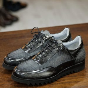 以下が含まれることがあります： グレーと黒の革製スニーカーのペア。アッパーにはテクスチャード加工が施されています。靴には黒い靴紐と厚い黒いソールが付いています。スニーカーは木製の表面に展示されています。