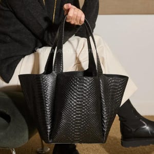 Op de afbeelding: Een zwarte tote bag met een slangenleertextuur wordt door een persoon vastgehouden. De tas heeft twee handvatten en een brede opening. Het ontwerp van de tas is donker en neutraal, en de textuur voegt visuele interesse toe.