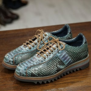 May include: A pair of textured, low-top sneakers with a snakeskin pattern in shades of teal, blue, and gray. The shoes have brown laces, a brown sole, and a ridged, dark gray outsole. The shoes are displayed on a wooden surface.