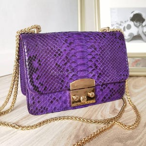 Puede incluir: Un vibrante bolso morado con un patrón de piel de serpiente. El bolso tiene un cierre dorado y una correa de cadena. El bolso rectangular parece ser de cuero, con unas dimensiones de aproximadamente 20 cm x 15 cm.