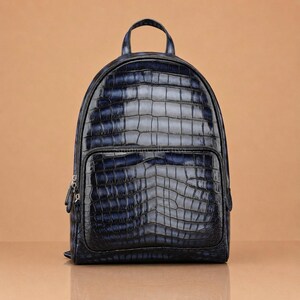 May include: A dark blue backpack featuring a glossy, embossed crocodile skin texture. The bag has a rounded top, a top handle, and a large front zippered pocket. The material displays a gradient of deep navy and black tones with a polished finish.