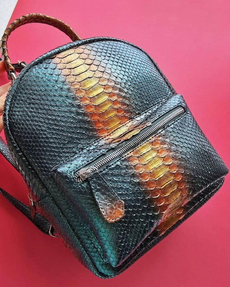 May include: A small, round backpack with a snakeskin pattern in shades of teal, gold, and brown. The backpack has a zippered front pocket and a braided handle. The zipper is silver-toned.