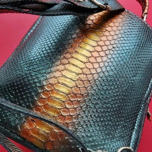 May include: A backpack featuring a snakeskin design in teal, gold, and brown hues. It has a top handle and adjustable straps. The metal hardware is silver. The background is a solid red.