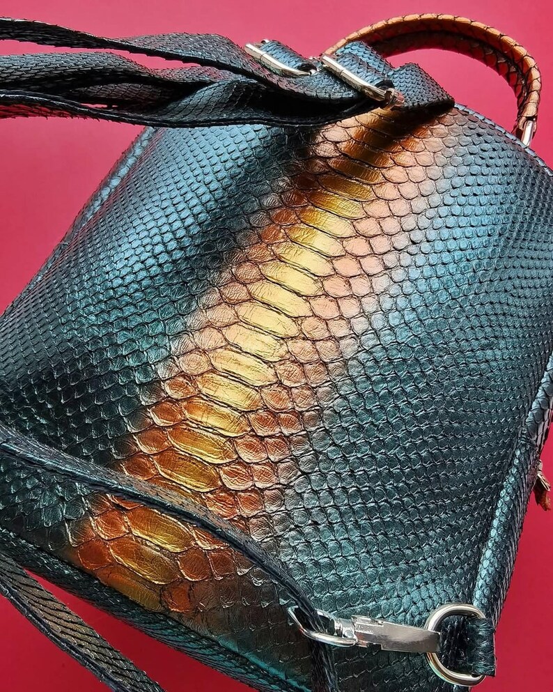 May include: A backpack featuring a snakeskin design in teal, gold, and brown hues. It has a top handle and adjustable straps. The metal hardware is silver. The background is a solid red.