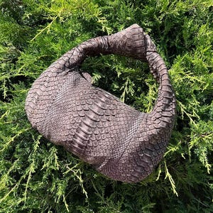 Peut inclure: Un sac à main gris avec un motif texturé et écailleux, ressemblant à de la peau de serpent. Le sac a une poignée incurvée et un corps arrondi. Le sac repose sur un lit de feuillage vert.